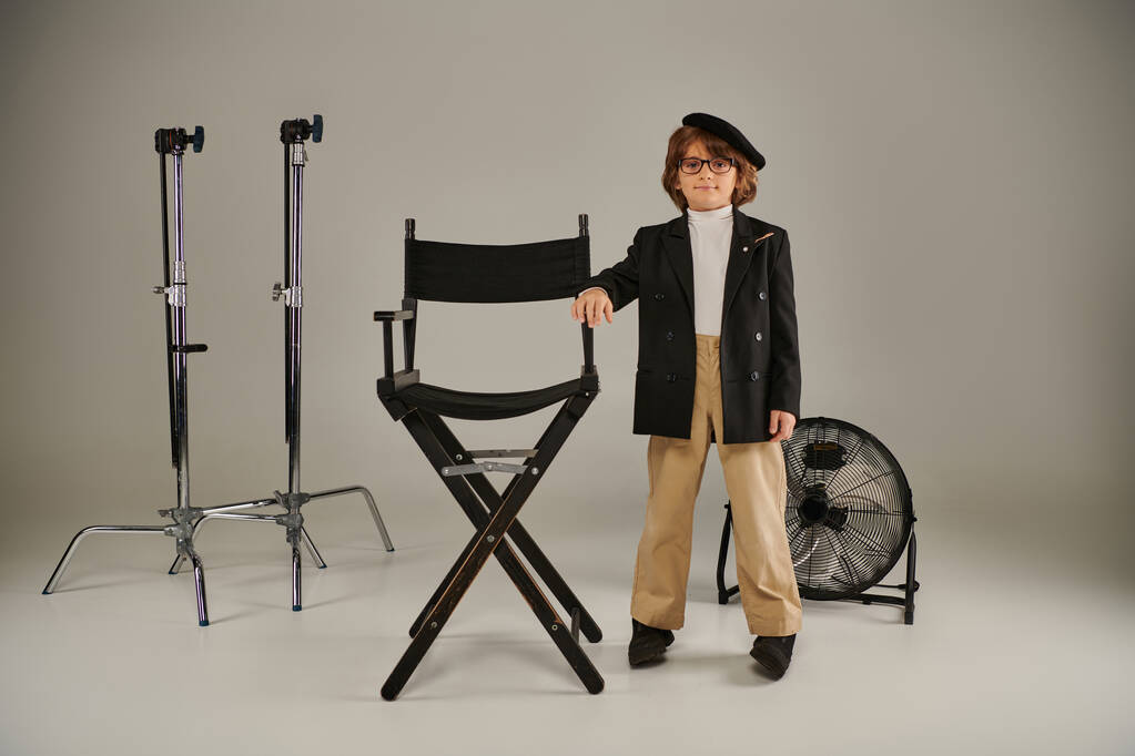 Netter Junge in Baskenmütze und schicker Freizeitkleidung steht selbstbewusst neben dem Regiestuhl auf grauem Hintergrund - Foto, Bild