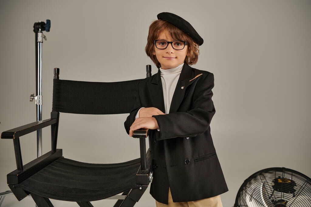 mignon garçon en béret et tenue élégante se tient avec confiance près de la chaise de directeur sur fond gris - Photo, image