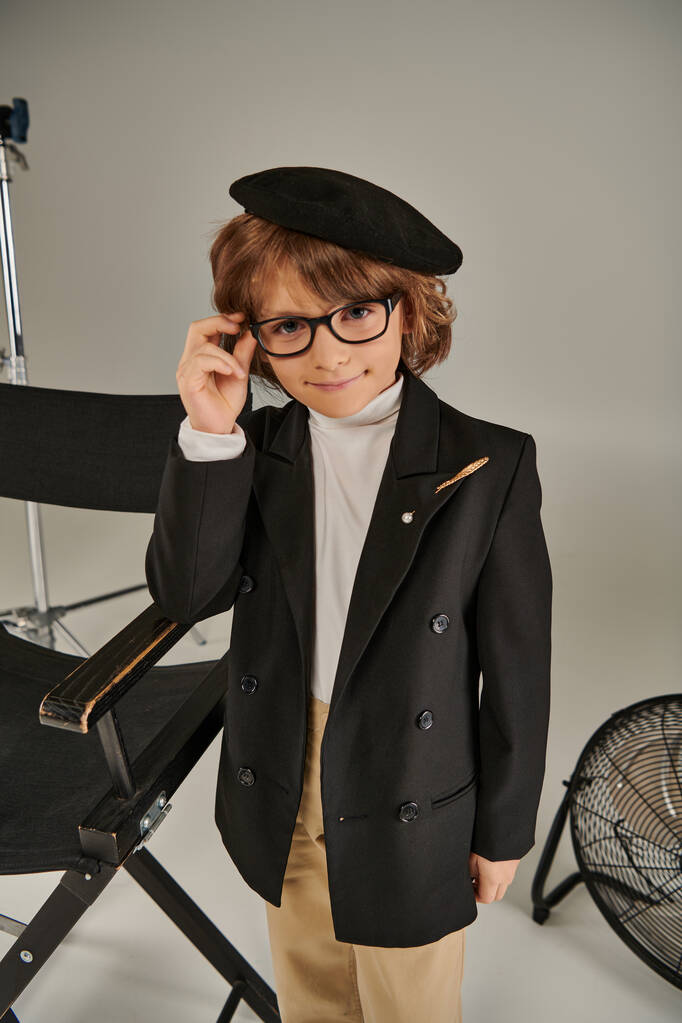 κομψό αγόρι φορώντας σακάκι και μπερέ, όπως ο ίδιος χαμογελά σε γκρι φόντο, παιδί ως σκηνοθέτης - Φωτογραφία, εικόνα