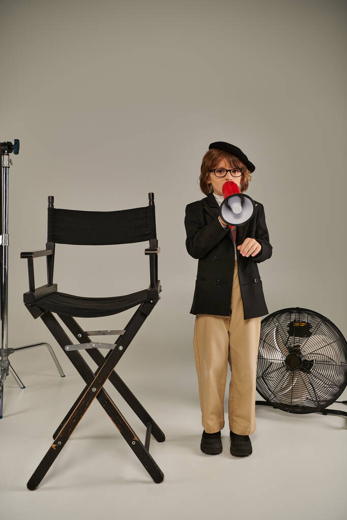 スタイリッシュな子供は,監督の椅子の近くに自信を持って立って,メガフォンで話す,灰色の背景 - 写真・画像