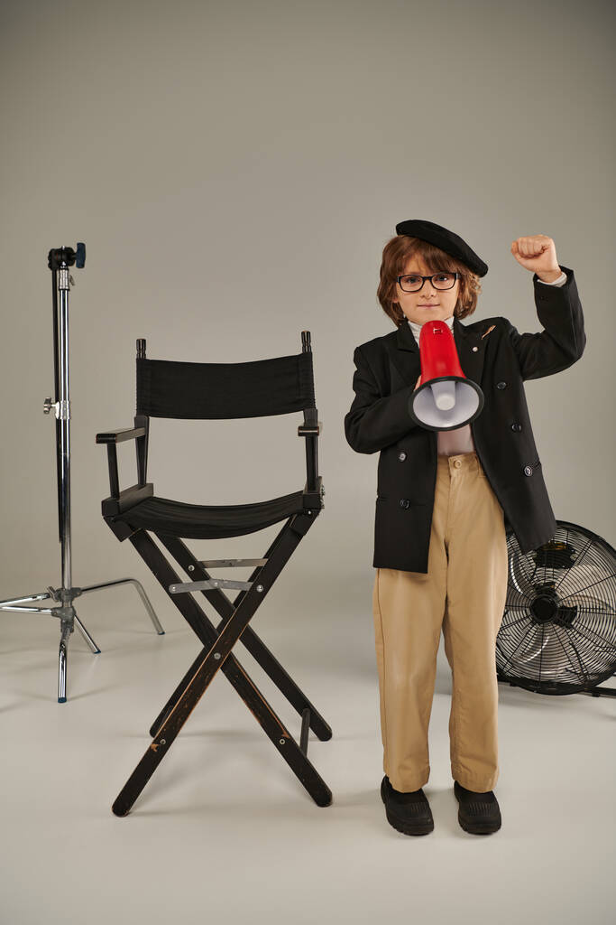 Młody aktywista w berecie stoi z megafonem i krzesłem reżyserskim na szarym, chłopiec jako filmowiec - Zdjęcie, obraz