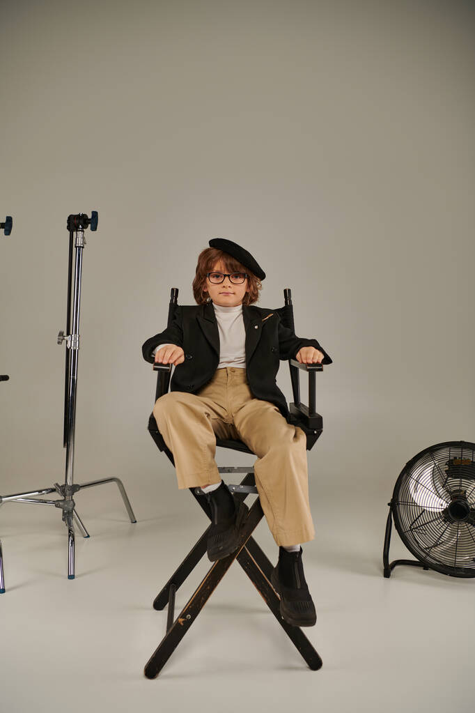 seguro cineasta chico en boina y gafas sentado en director silla en gris telón de fondo - Foto, imagen