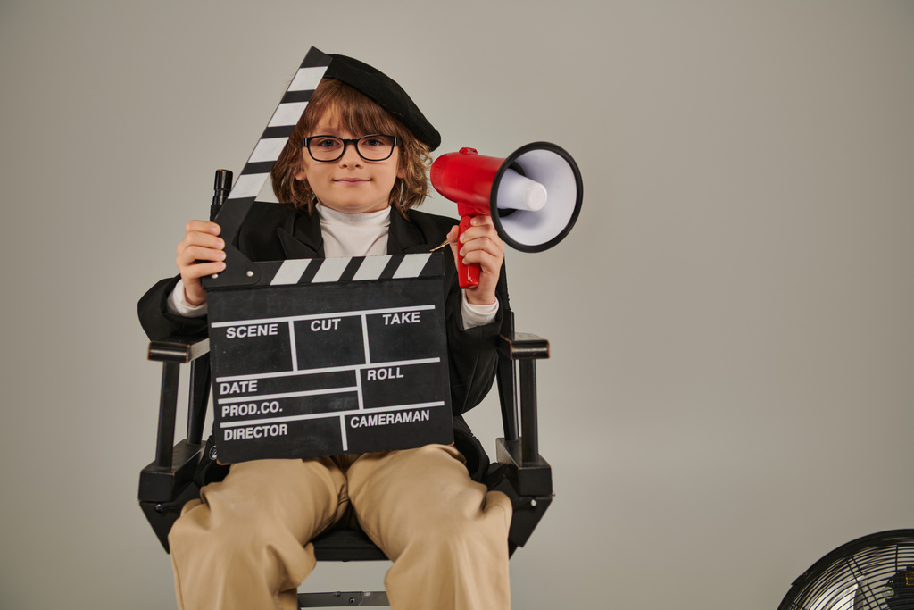 мальчик в берете и очках, держащий доску объявлений и красный мегафон, сидящий на режиссерском стуле - Фото, изображение