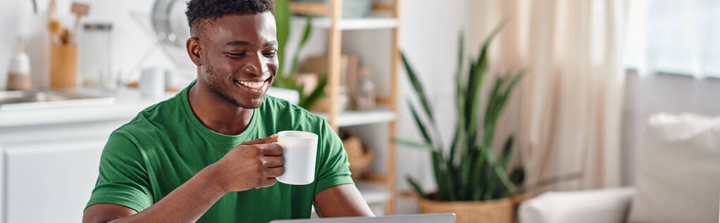 Uśmiechnięty Afroamerykanin, cieszący się filiżanką kawy i uśmiechnięty w kuchni, poziomy baner - Zdjęcie, obraz