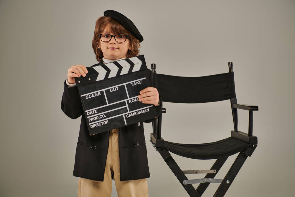 stylish filmmaker boy in beret and glasses holding clapper board and standing near director chair - Photo, Image