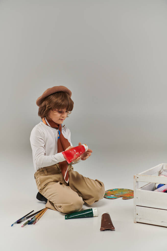 мальчик преклоняет колени на полу, окруженный красками в трубах и деревянной коробкой для инструментов, молодой художник в берете - Фото, изображение