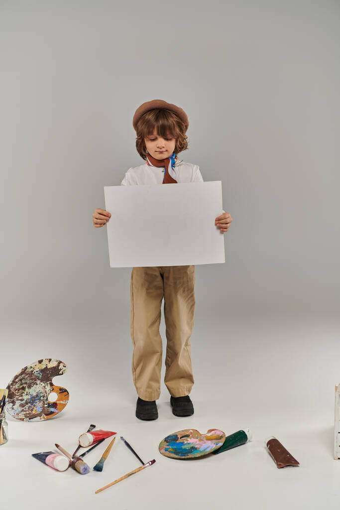 jovem artista segurando papel em branco perto de pincéis de pintura e paleta no chão, menino em boina e cachecol - Foto, Imagem