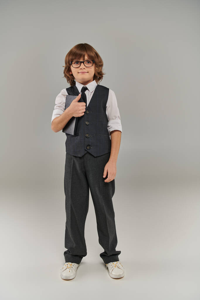 A young boy dressed in a fashionable vest and tie holding book and standing on grey background - Photo, Image