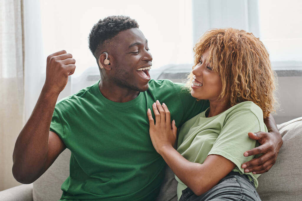 pareja afroamericana emocionada abrazando, hombre negro sordo en audífono y mujer en aparatos ortopédicos - Foto, imagen
