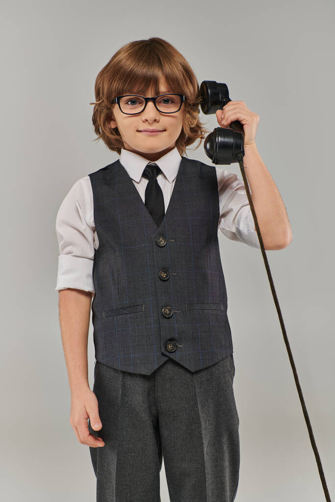 A well-dressed young gentleman in elegant vest with tie and glasses holding a telephone on grey - Photo, Image