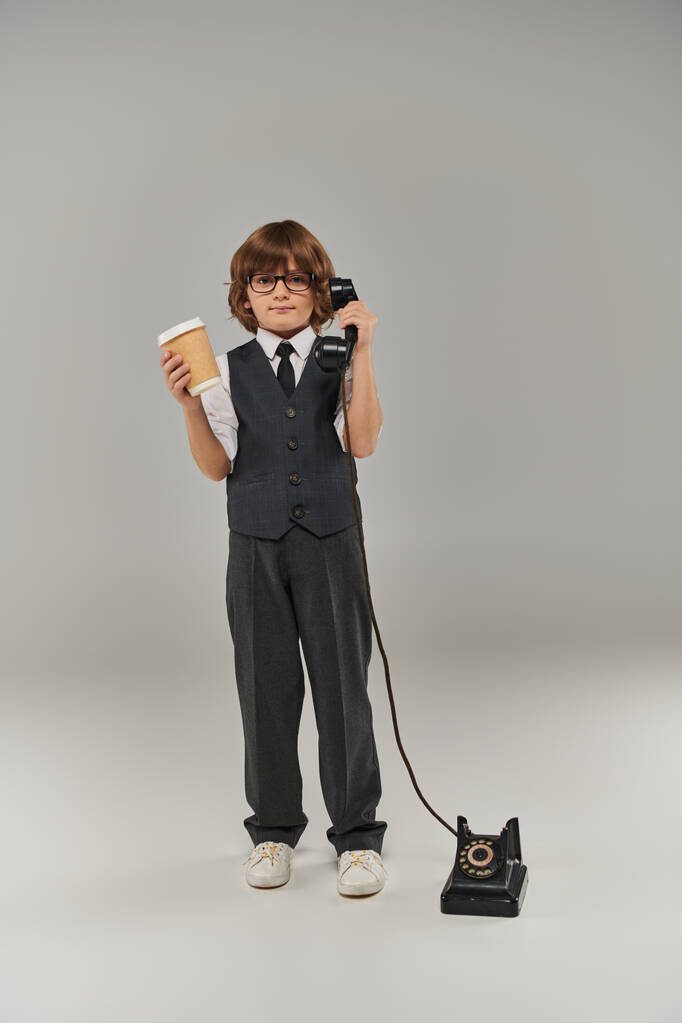 menino em óculos e elegante traje formal segurando telefone retro e beber em copo de papel em cinza - Foto, Imagem