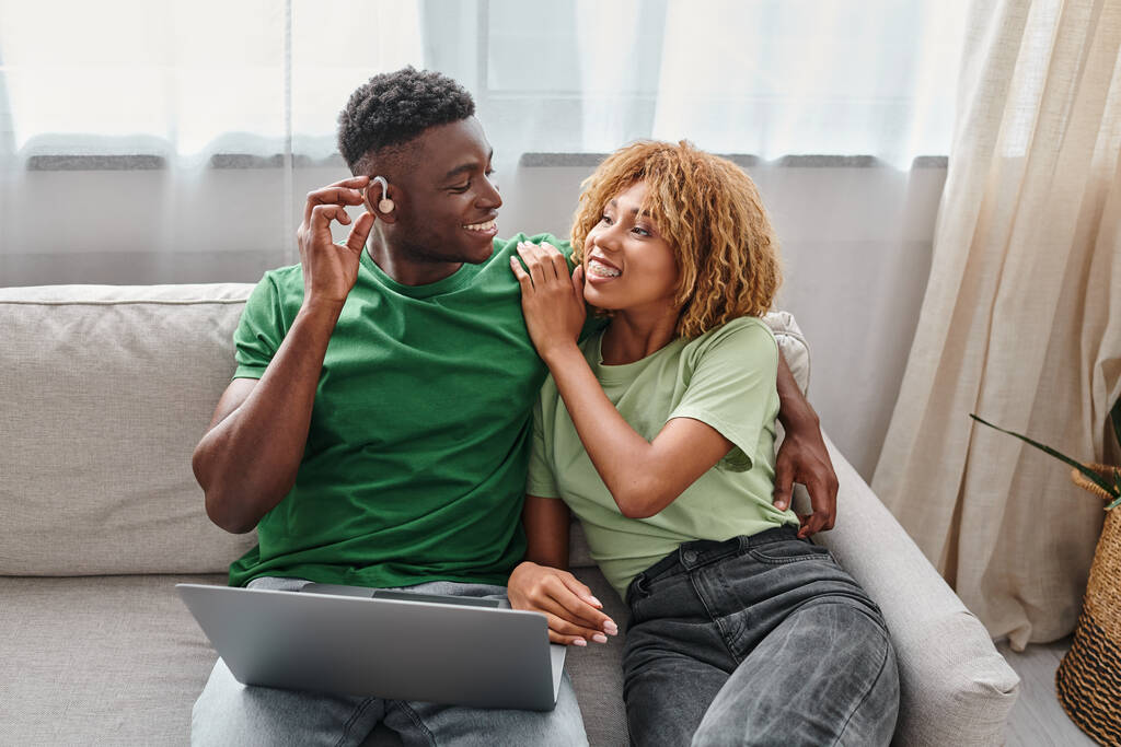 felice uomo africano americano in apparecchio acustico seduto sul divano con la fidanzata e utilizzando il computer portatile - Foto, immagini