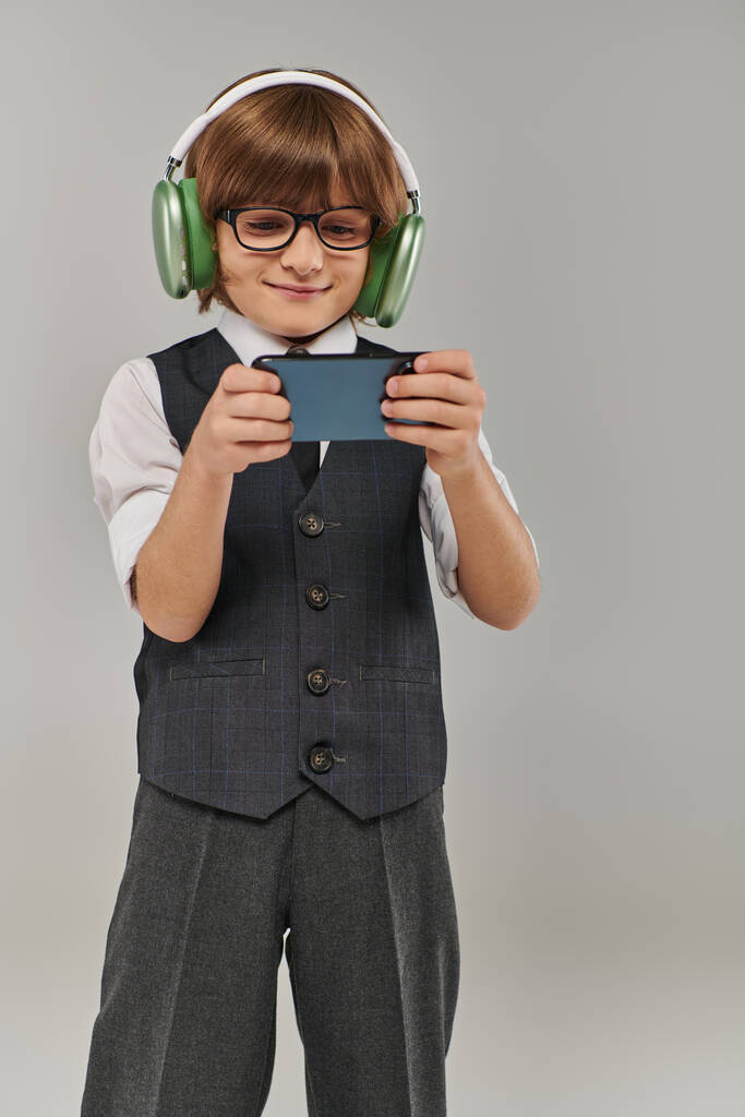 menino elegante em óculos e traje elegante ouvir música em fones de ouvido e jogar jogo móvel - Foto, Imagem