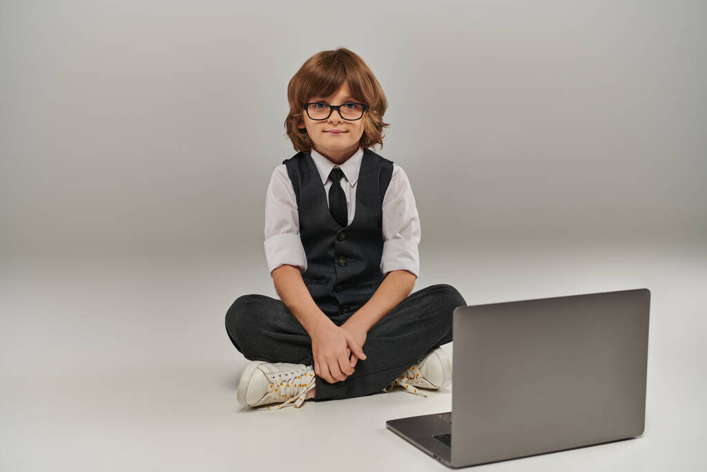 garçon en tenue élégante avec gilet et pantalon assis près d'un ordinateur portable sur gris, futur homme d'affaires - Photo, image