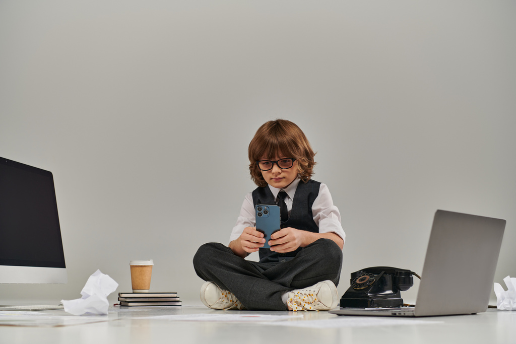 Junge sitzt auf dem Boden, vertieft in die Technologie, wie er sowohl Telefon als auch Laptop, zukünftiger Geschäftsmann - Foto, Bild