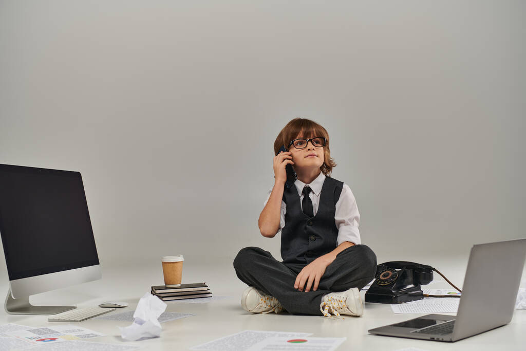 メガネとフォーマルな服装の男の子がスマートフォンで話し,オフィス機器に囲まれて座っている - 写真・画像