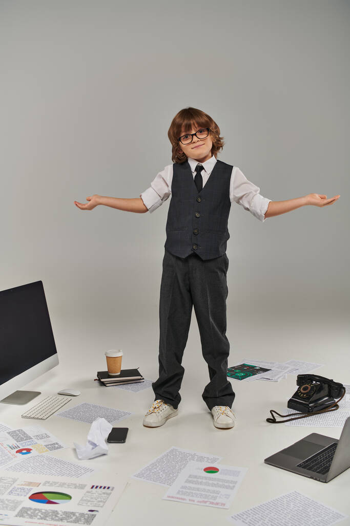 bambino confuso in occhiali e abbigliamento formale circondato da attrezzature e dispositivi per ufficio in piedi sul grigio - Foto, immagini