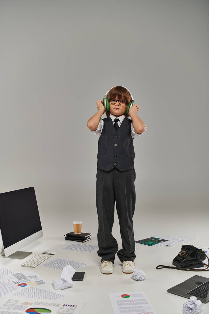 αγόρι βυθίζεται στον ψηφιακό κόσμο, παιδί με ακουστικά στέκεται κοντά σε συσκευές και χαρτιά στο πάτωμα - Φωτογραφία, εικόνα