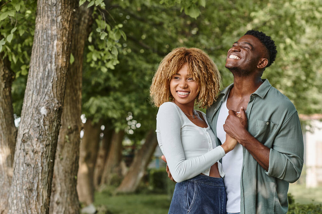 joyeux couple afro-américain tenant la main et s'embrassant à l'extérieur, femme en bretelles - Photo, image