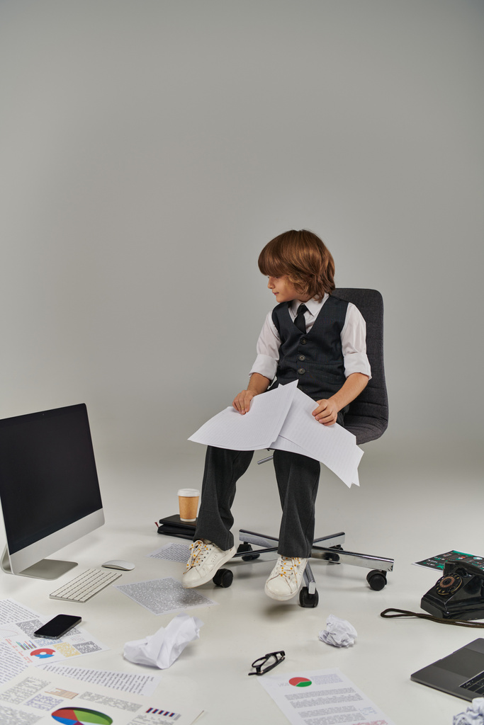 Niño en traje formal revisa documentos rodeados de suministros de oficina, futuro profesional - Foto, imagen