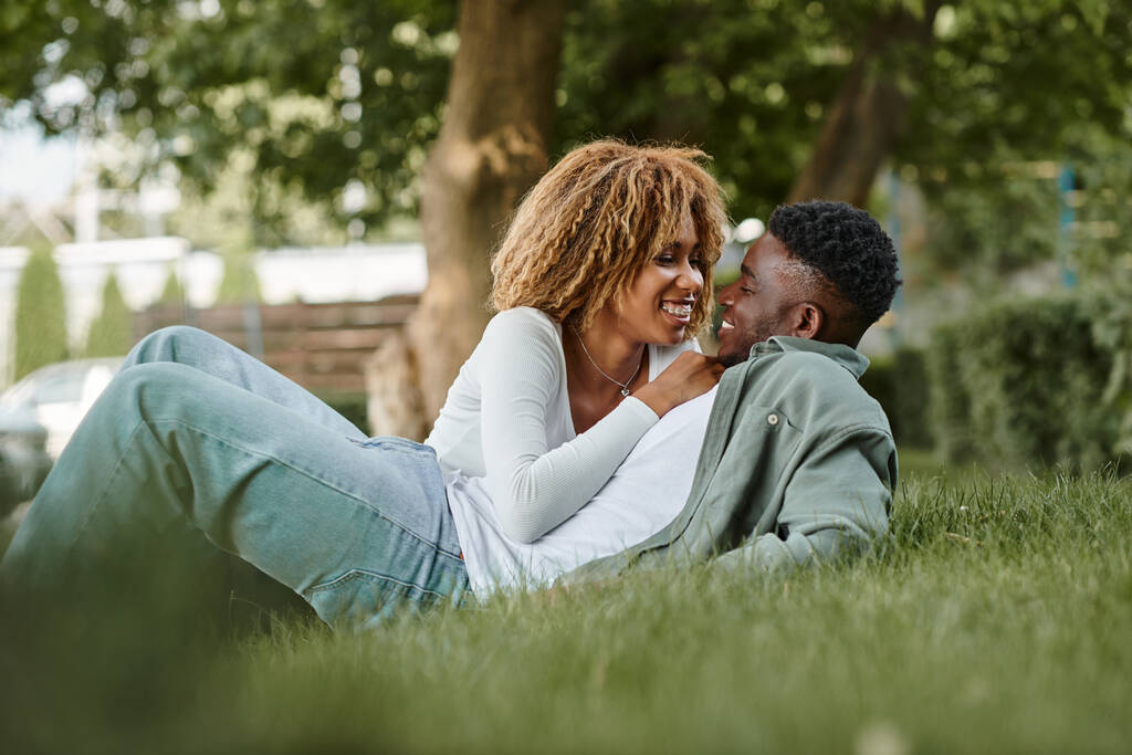 χαρούμενο νεαρό αφρικανικό ζευγάρι που μοιράζεται μια τρυφερή ματιά ενώ κάθεται σε ένα γρασίδι στο πάρκο - Φωτογραφία, εικόνα