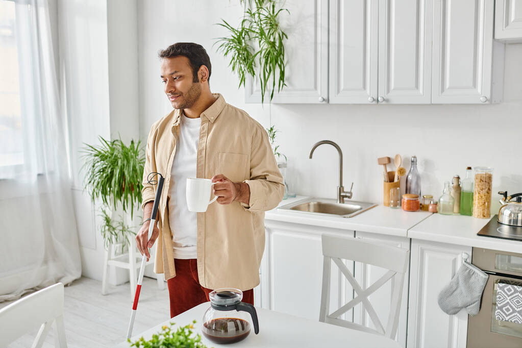 knappe gehandicapte indiaanse man in gezellige huiskledij met wandelstok met koffiebeker op keuken - Foto, afbeelding