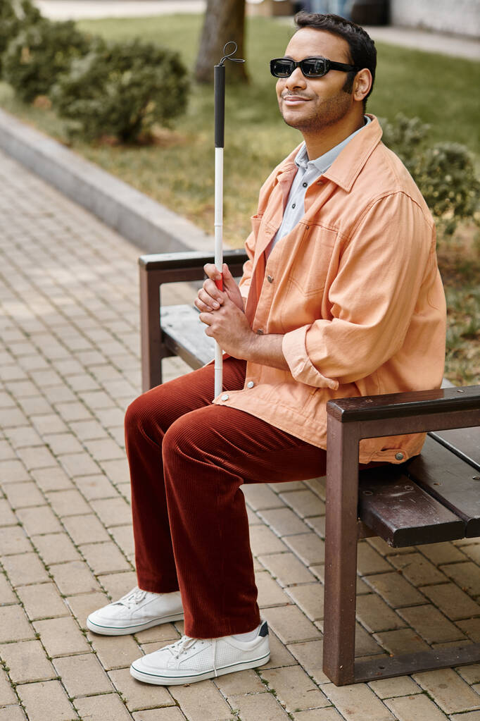 χαρούμενος ινδός τυφλός με πορτοκαλί σακάκι κάθεται έξω στον πάγκο με το μπαστούνι και τα γυαλιά - Φωτογραφία, εικόνα