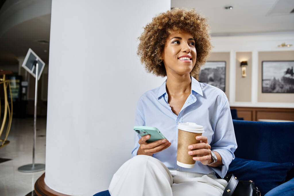 mujer afroamericana rizada feliz con café para llevar y el uso de teléfono inteligente en el vestíbulo del hotel - Foto, imagen