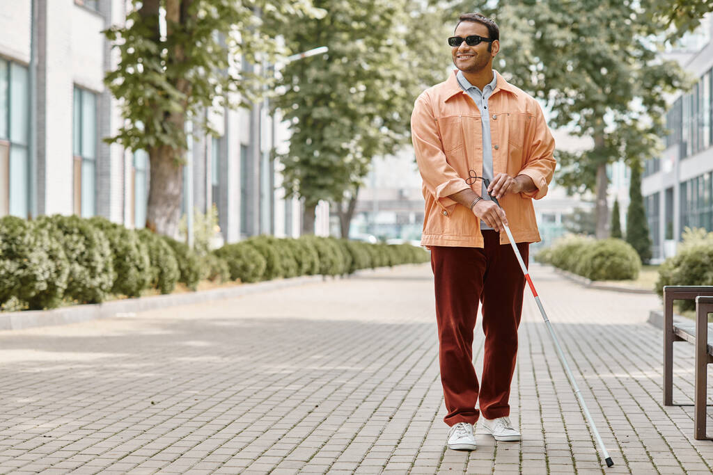 alegre indio ciego con gafas y bastón en naranja chaqueta viva caminando en el parque - Foto, Imagen