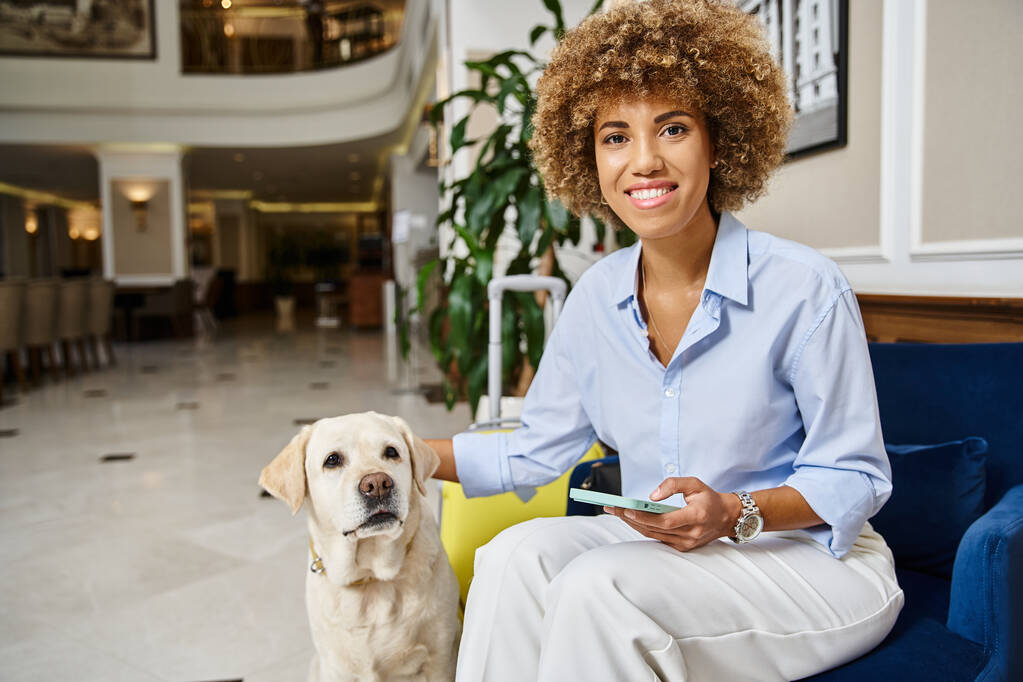 viajero feliz con labrador y teléfono en un hotel que acepta mascotas, perro y mujer afroamericana - Foto, imagen