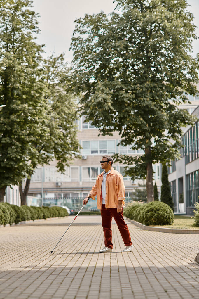 vreugdevolle indiaanse blinde man in oranje jasje met bril en wandelstok wandelen in park - Foto, afbeelding
