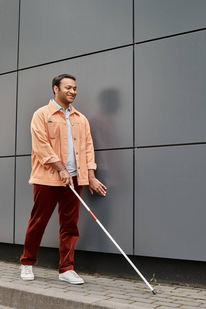 joyeux aveugle indien en veste orange avec bâton d'aide marchant avec mur gris sur fond - Photo, image