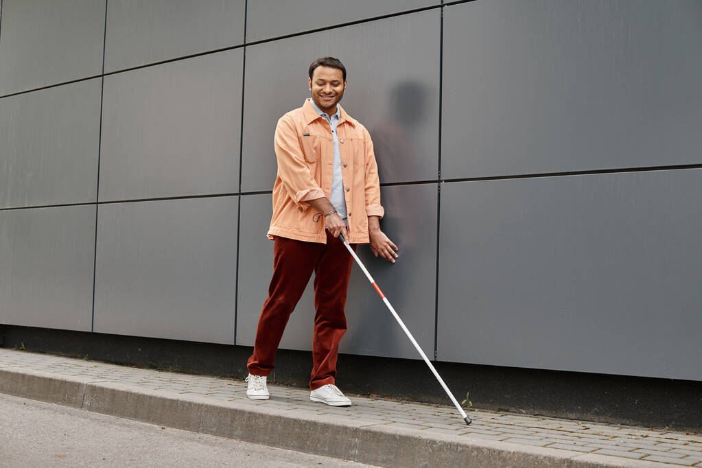 alegre hombre ciego indio en chaqueta naranja con ayuda palo caminando con la pared gris en el telón de fondo - Foto, imagen