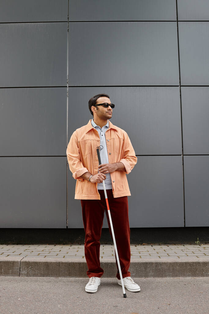 dobře vypadající indický zdravotně postižený muž v oranžové bundě s brýlemi a hůl s šedou stěnou na pozadí - Fotografie, Obrázek