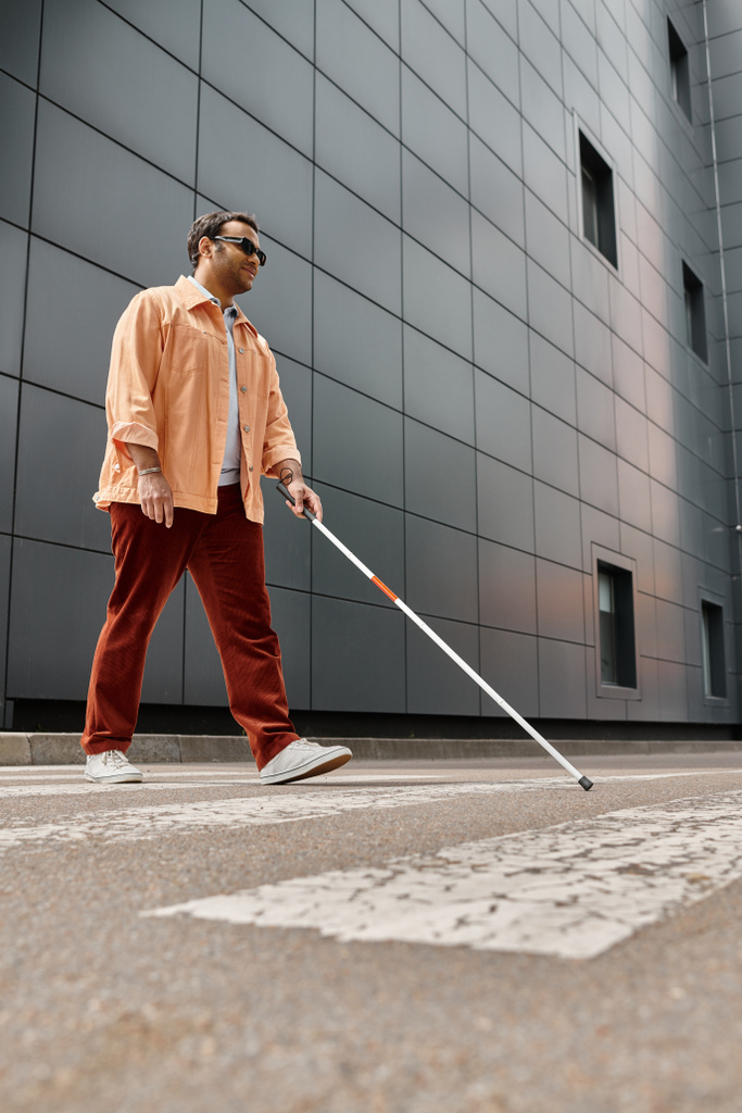 upbeat indian blind man in orange jacket with walking stick and glasses with gray wall on backdrop - Photo, Image