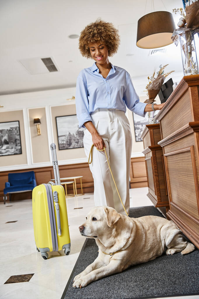 alegre mujer afroamericana con su labrador registrándose en un hotel que acepta mascotas en recepción - Foto, Imagen