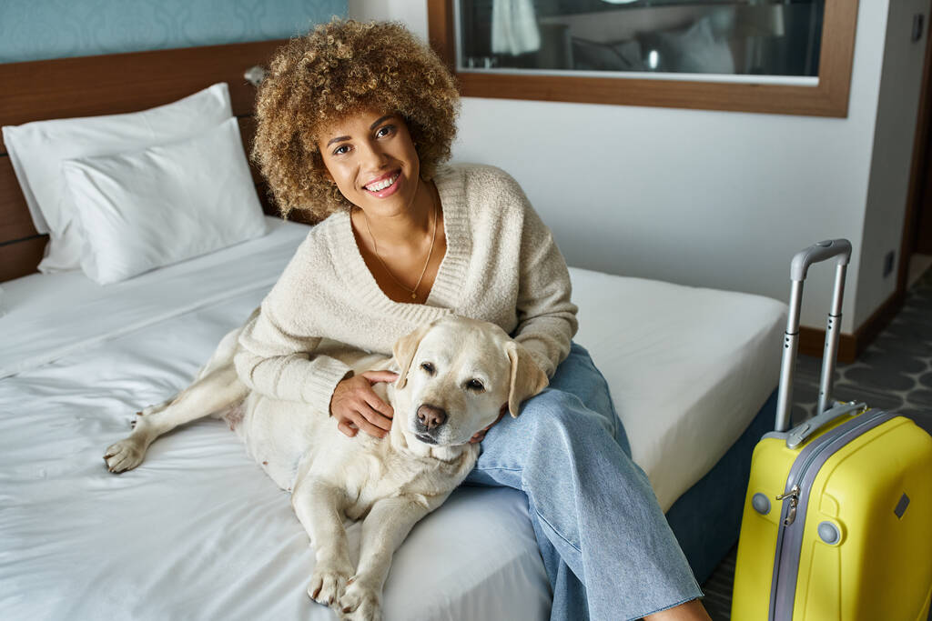 happy african american woman cuddling labrador near luggage in pet-friendly hotel room - Photo, Image