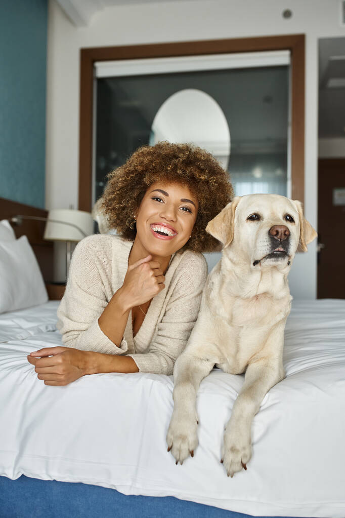 alegre mujer afroamericana con su labrador acostado en una cama en un hotel que acepta mascotas, viajar - Foto, Imagen
