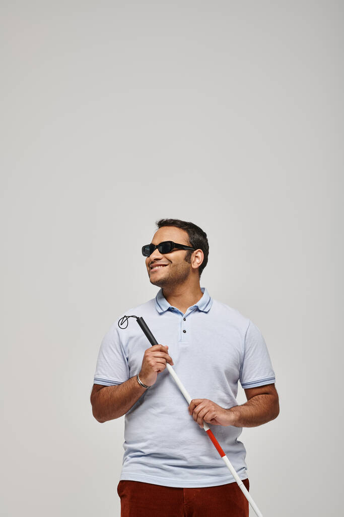 gioioso cieco indiano in maglietta blu con occhiali e bastone da passeggio in posa su sfondo grigio - Foto, immagini