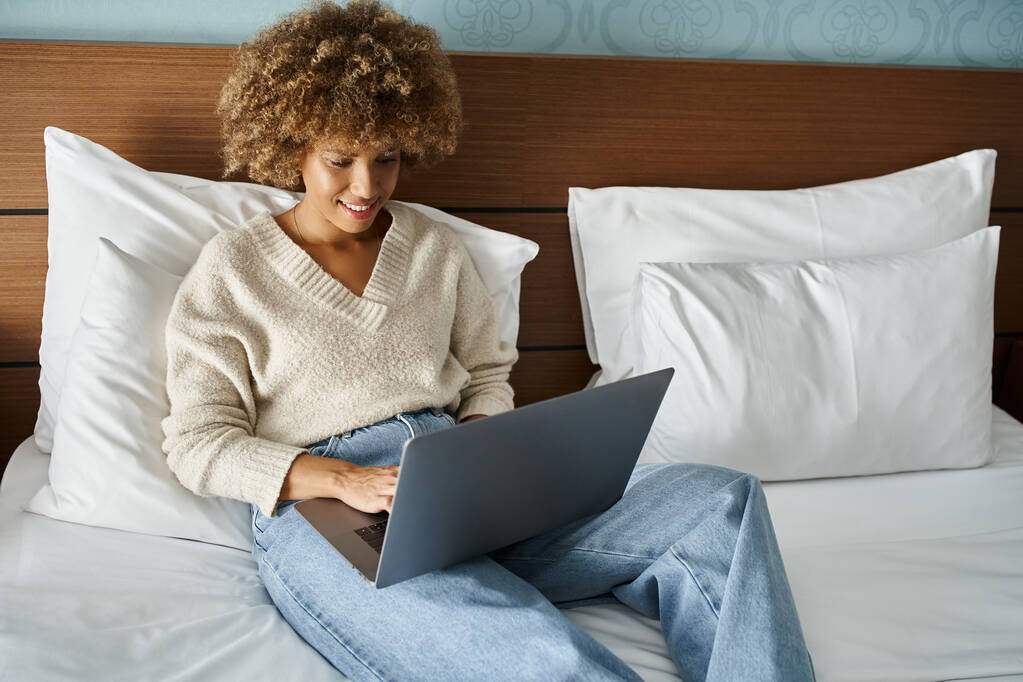 mujer afroamericana feliz y rizada sonriendo mientras trabaja en el ordenador portátil en la cama en una habitación de hotel - Foto, imagen