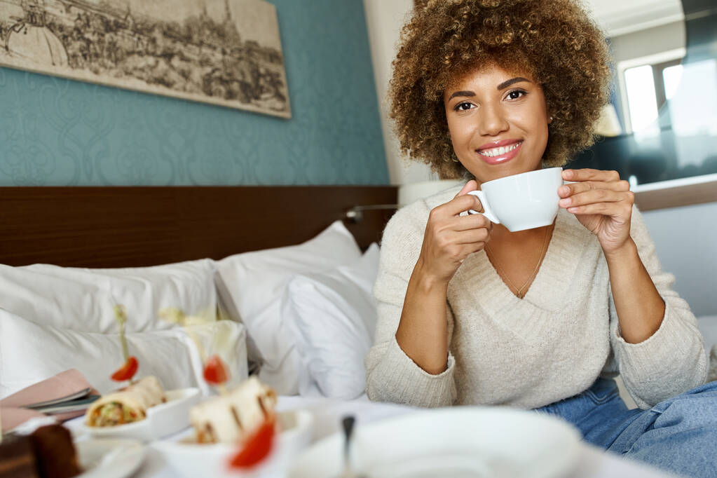 vreugdevolle Afrikaans-Amerikaanse vrouw die beker vasthoudt en op bed zit in hotelkamer, roomservice en comfort - Foto, afbeelding