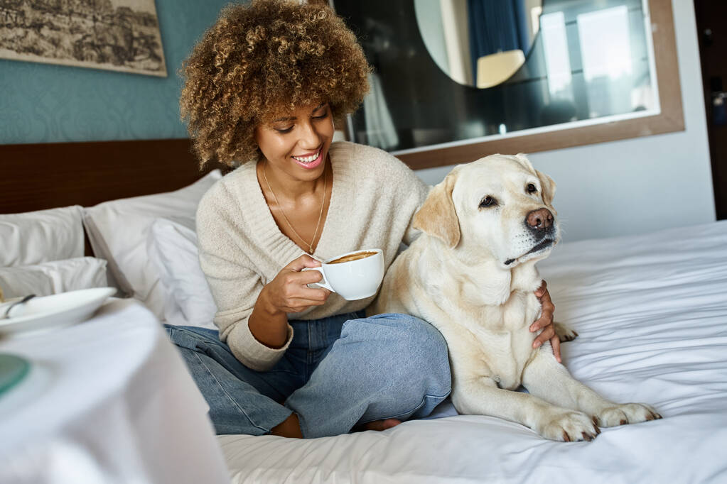 alegre africana americana mujer celebración taza y abrazo su labrador perro en mascota friendly hotel habitación - Foto, imagen