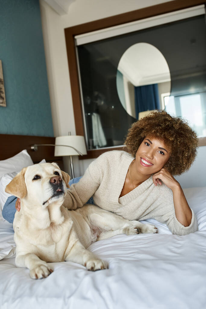 молодая африканская американка, лежащая на кровати и обнимающая свою собаку-лабрадора в отеле для животных - Фото, изображение