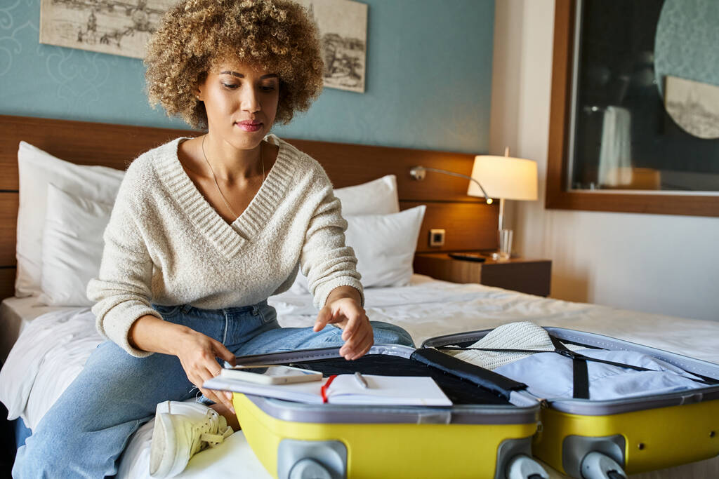 νεαρή Αφροαμερικανή γυναίκα με σγουρά μαλλιά ξεπακετάρει τις αποσκευές της στο δωμάτιο του ξενοδοχείου, έννοια απόδραση - Φωτογραφία, εικόνα