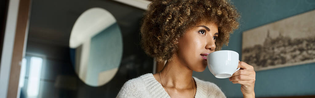 mujer afroamericana de pelo rizado bebiendo café mientras mira hacia otro lado en la habitación de hotel, banner de viaje - Foto, imagen
