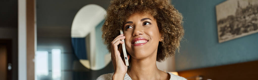 Wesoła Afroamerykanka z kręconymi włosami podczas rozmowy telefonicznej w pokoju hotelowym, baner horyzontalny - Zdjęcie, obraz