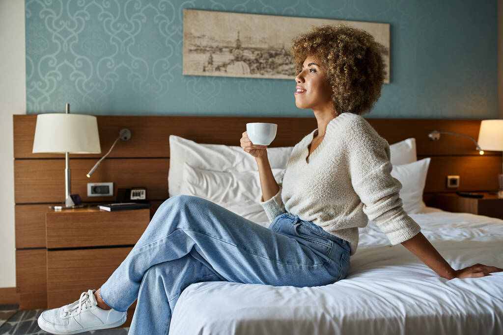 happy and young african american woman enjoying coffee while sitting on hotel bed, weekend getaway - Photo, Image