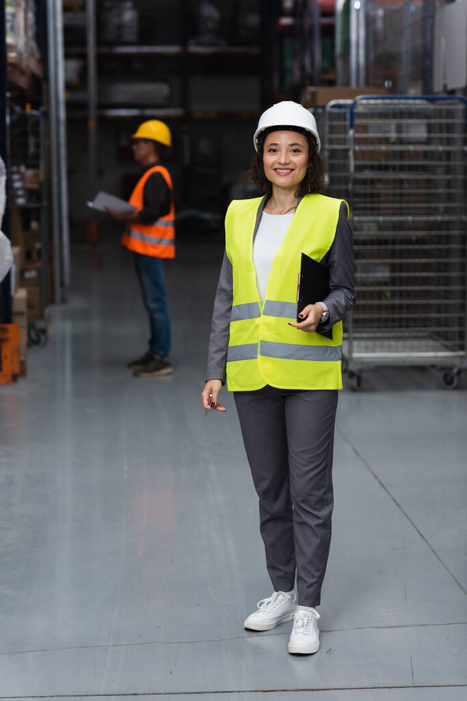 happy warehouse manager in safety vest and hard hat holding clipboard, logistics and distribution - Photo, Image