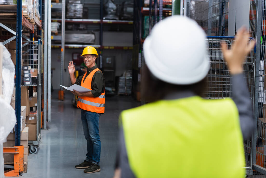 happy supervisor in hard hat waving at colleague while greeting each other in warehouse - Photo, Image