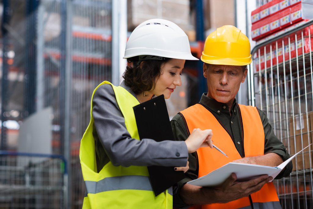 middle aged warehouse supervisor in safety vest showing paperwork to his female employee with pen - Photo, Image